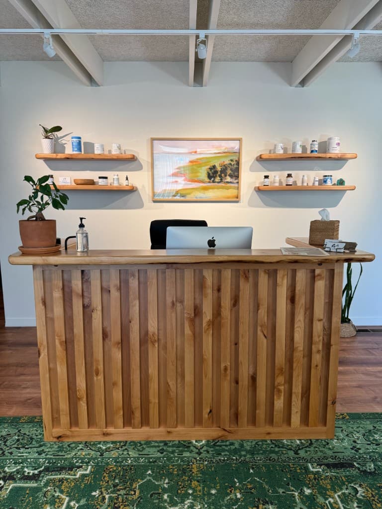 Light wood reception desk with live-edge counter, computer, and coastal artwork at Coastal Chiropractic SLO